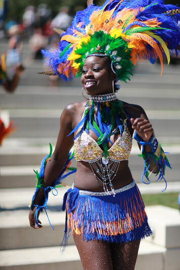 A Toronto Caribbean Carnival participant wearing a blue, yellow, orange, and green costume with elaborate beading and feather details as she walks confidently down the street, a smile on her face