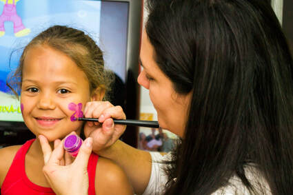 A woman painting a child's face. The child is very excited, and a purple and pink flower is being painted on her cheek. 