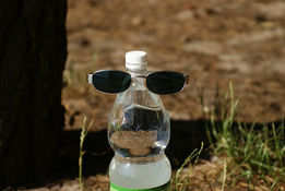 A single-use plastic water bottle sitting in the middle of a field with a pair of black-tinted sunglasses resting on it. 