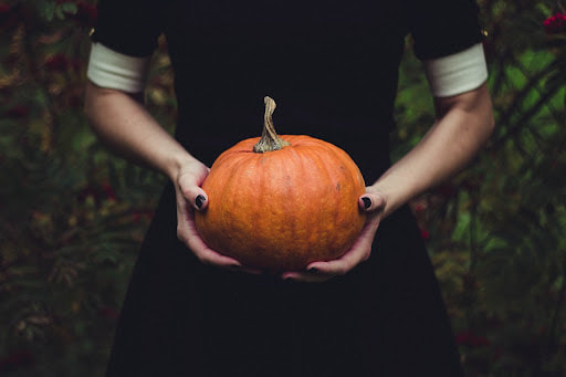 Person in a black dress with white sleeve cuffs wearing black nail polish and holding a small orange pumpkin