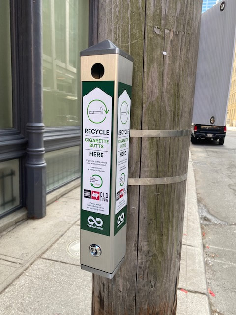 A slim metal cigarette butt recycling receptacle with a green and white sticker explaining the purpose of the bin and which partners were involved in the project.